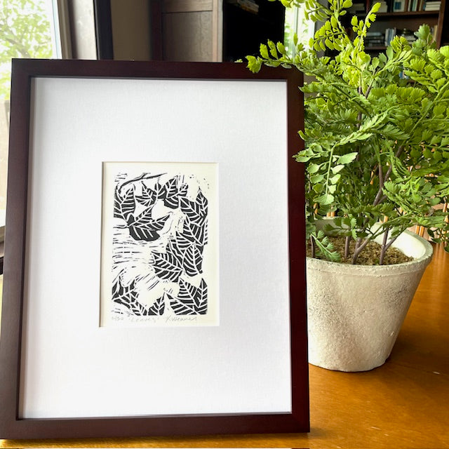 Fall leaf linocut print by Katie Weaver in black ink displayed on a table beside a plant 